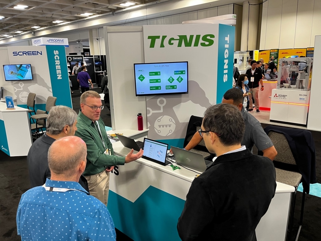 A booth at a trade fair for the company Tignis with four men standing around a computer while a fifth man explains what is on the screne.