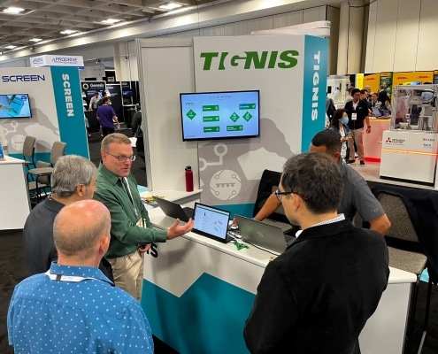 A booth at a trade fair for the company Tignis with four men standing around a computer while a fifth man explains what is on the screne.