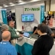 A booth at a trade fair for the company Tignis with four men standing around a computer while a fifth man explains what is on the screne.