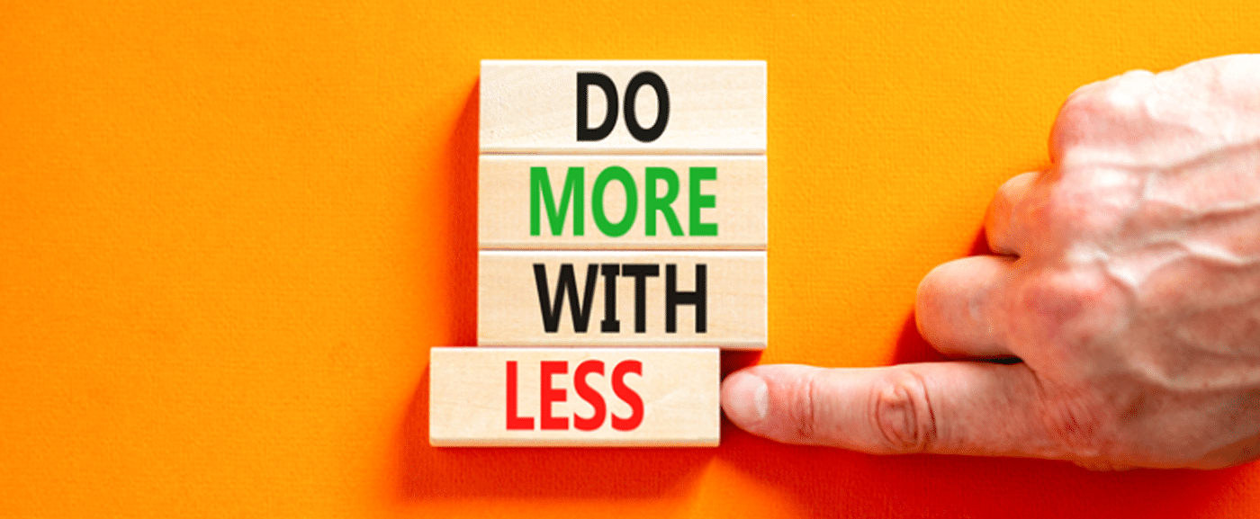 domorewithless_1400 An orange background with four wooden blocks arranged vertically. Each one reads one word 'Do' 'More' 'With' 'Less' and there is a hand pushing the last block 'Less' out of alignment with the others